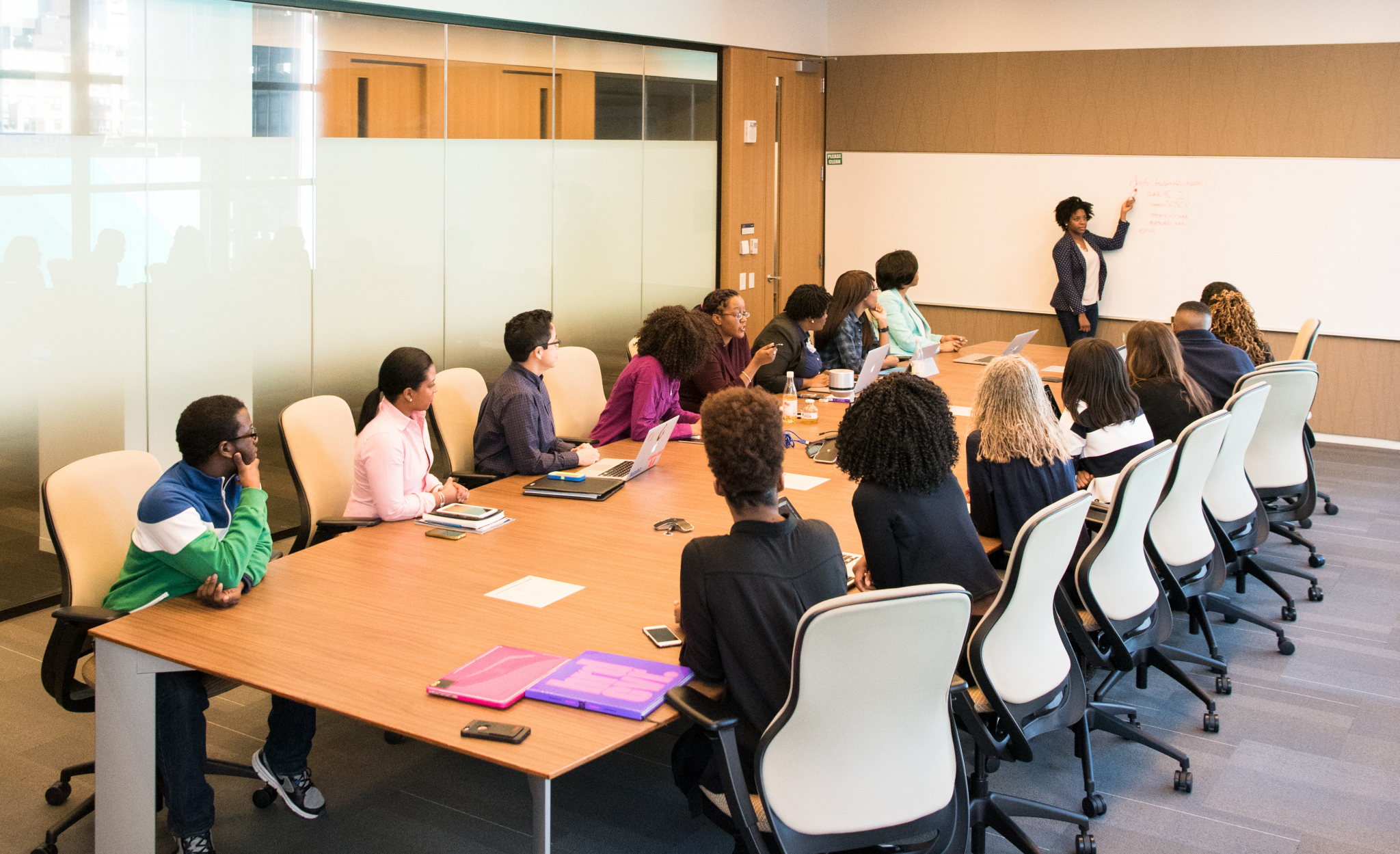 Conference Room with diverse people sitting at long table watching a woman at the white board